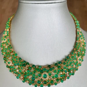 The image showcases a lavish necklace adorned with numerous green emerald gemstones and sparkling white diamonds, set in what appears to be yellow gold. The necklace is displayed on a gray mannequin bust, highlighting its intricate design and luxurious appeal.