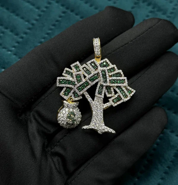 This image shows a luxurious pendant held against a black gloved hand. The pendant is shaped like a stylized tree, with branches filled with green and white sparkling stones. Hanging from one side of the tree is a small money bag charm, also fully encrusted with shimmering stones, symbolizing wealth and prosperity. The entire piece is crafted in 925 sterling silver, with parts of it finished in a gold tone, particularly around the bail (the loop where the chain goes through) and the money bag. The pendant has a bold, flashy, hip hop aesthetic, emphasizing luxury, success, and high fashion.