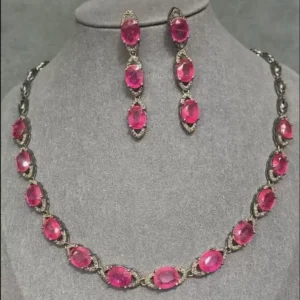 The image showcases a vintage-style necklace and matching drop earrings. Both pieces feature oval-cut, vibrant pink crystals bezel-set in what appears to be sterling silver 925. The design evokes an Art Deco aesthetic with its symmetrical arrangement and clean lines. The set is displayed on a gray mannequin bust.